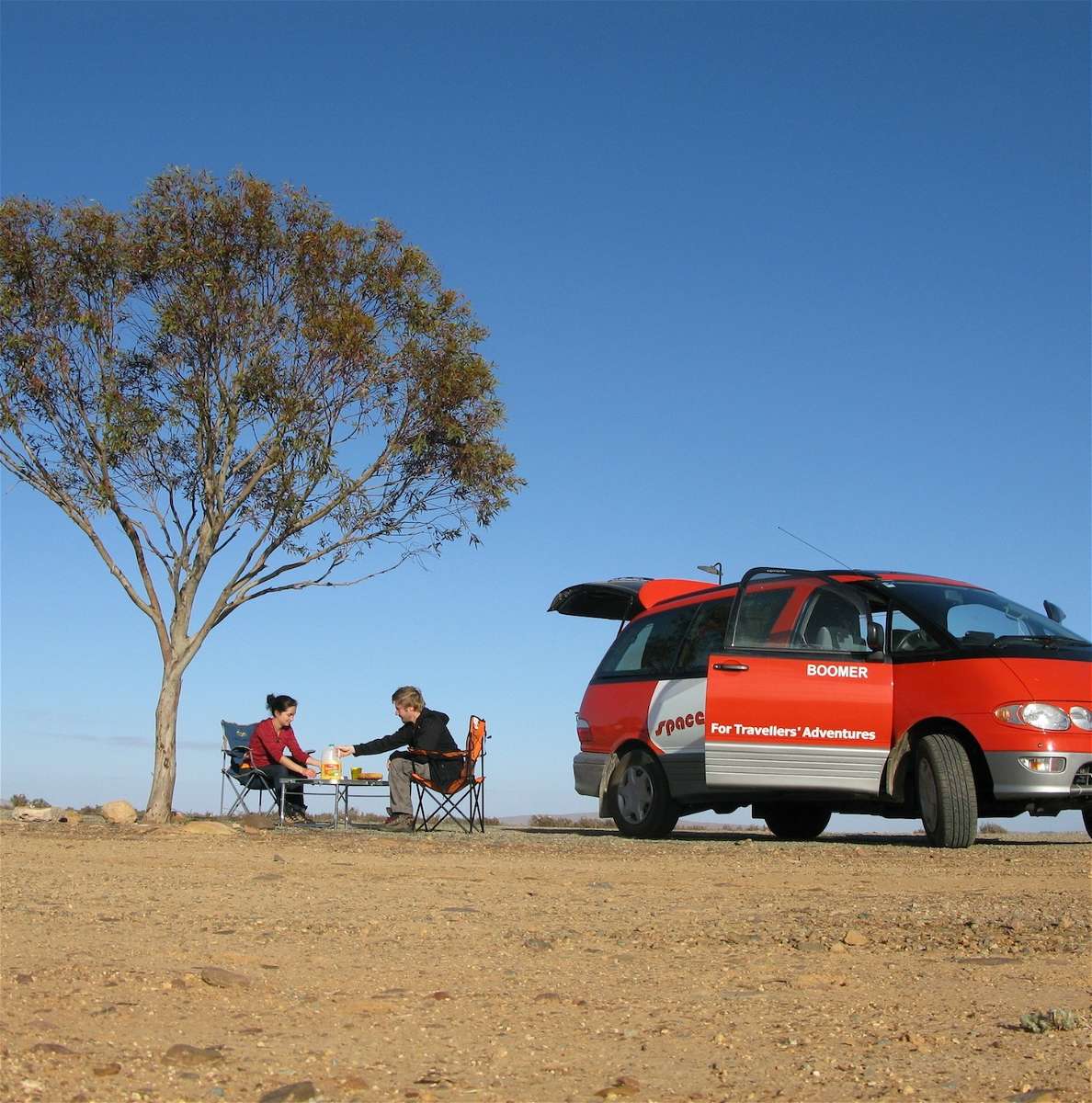 Campervan Hire Australia - red campervan with couple sitting at a table under a tree