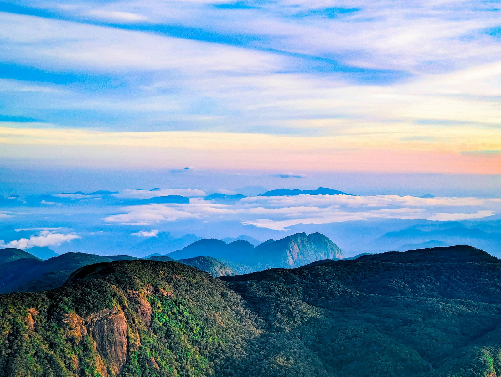 Sunrise at the top of a mountain peak overlooking misty jade green valleys with pale clouds and a rosey blue sky
