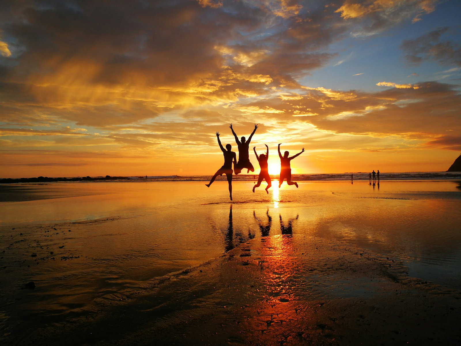 Four travellers jumping in the air at a burnt orange sunset on a beach when the tide is low, some more people in distance