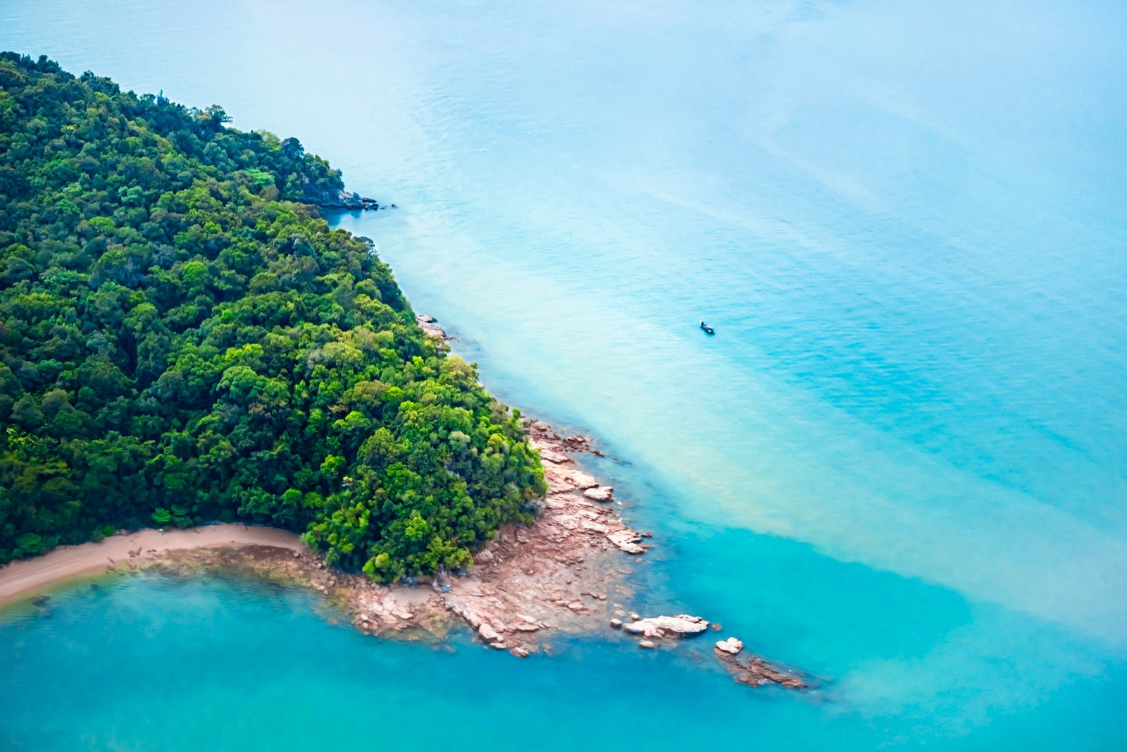 Aerial shot of secluded island with pink-tinted rocks, bright blue seas and emerald shrubbery with single ship in distance