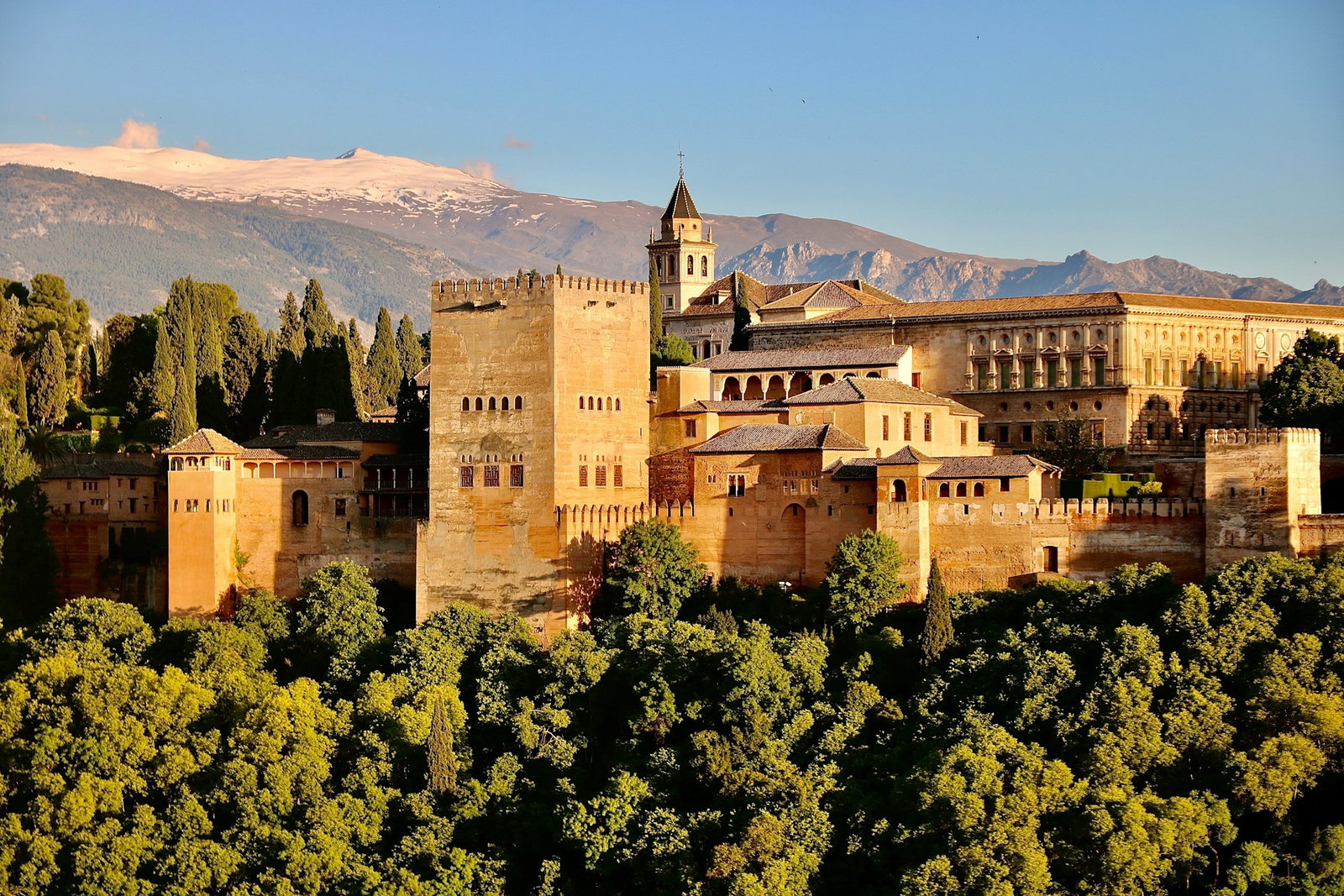 Alhambra castle with golden architecture nestled into hilltops with trees below and mountains behind on clear day