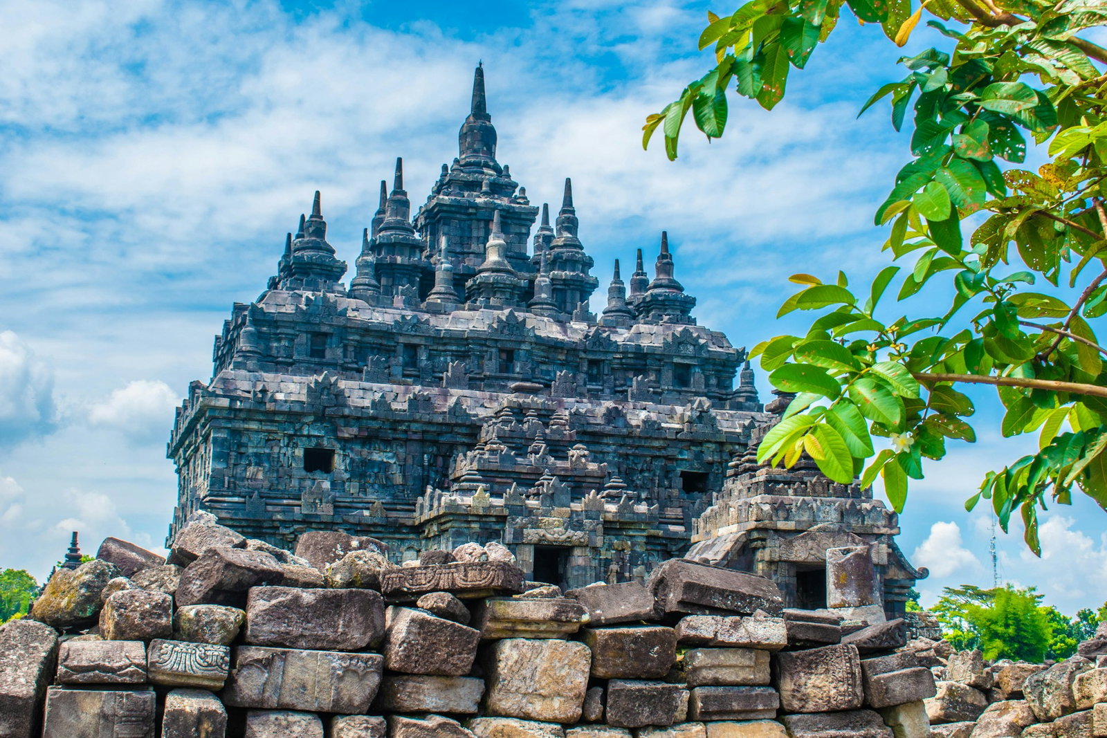 Ancient temple with ornate pagodas at the top and brick wall in front on a cloudy blue sky day with a tree to the right