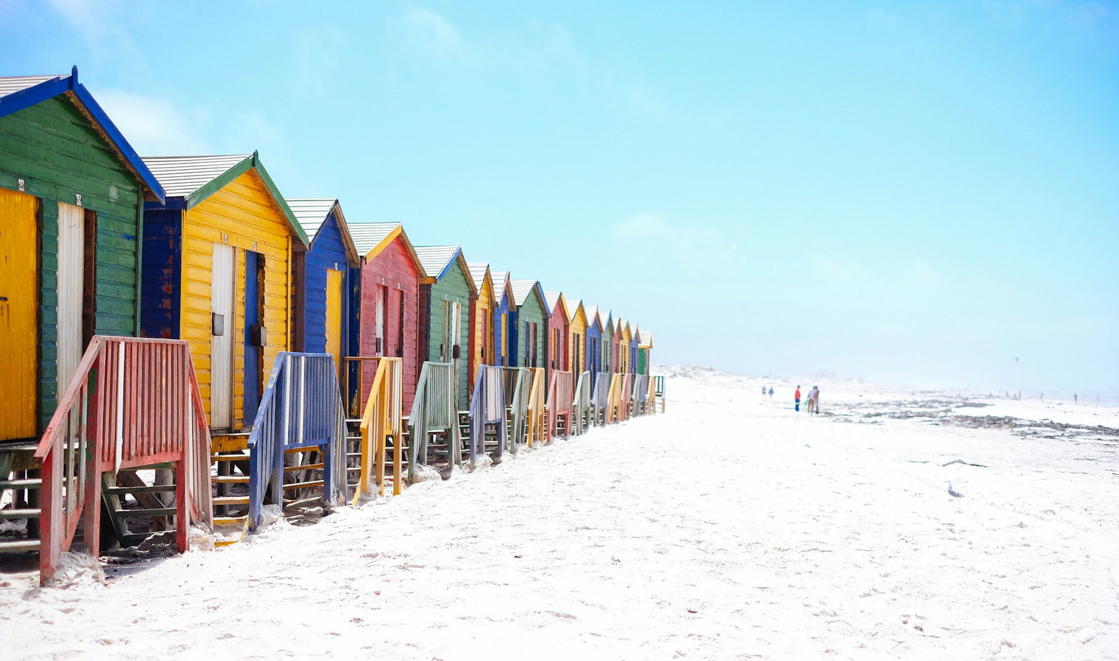 Row of vibrant huts to the left on white sand beach with pale blue sky and people in the distance, Cape Town