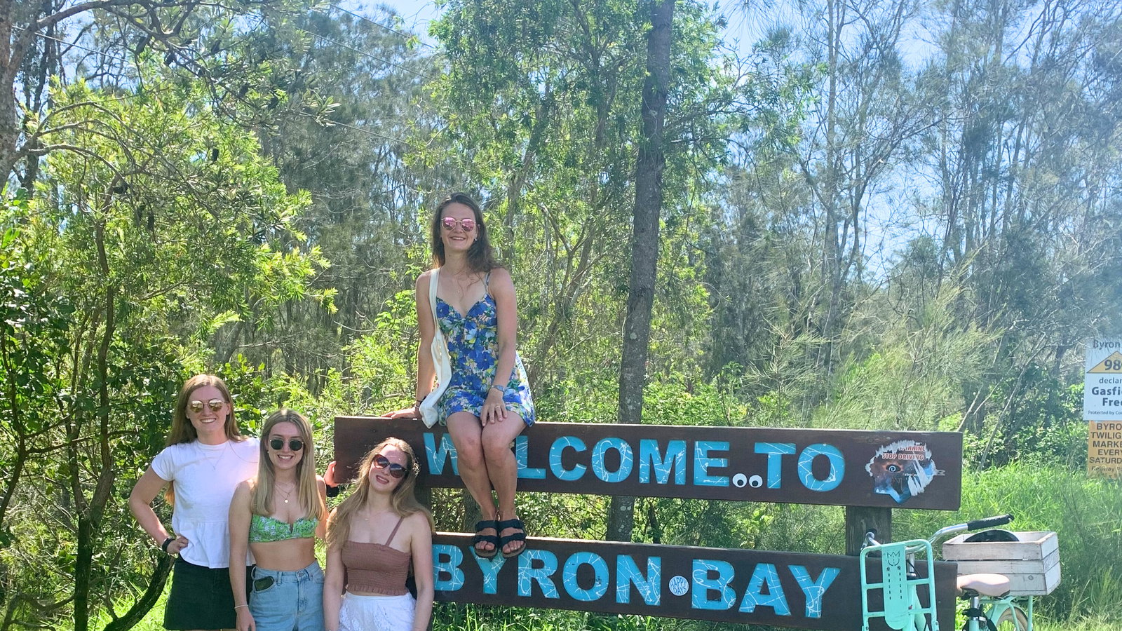 Group of travellers with a bike at 'Welcome to Byron Bay' Sign, Byron Bay, Australia