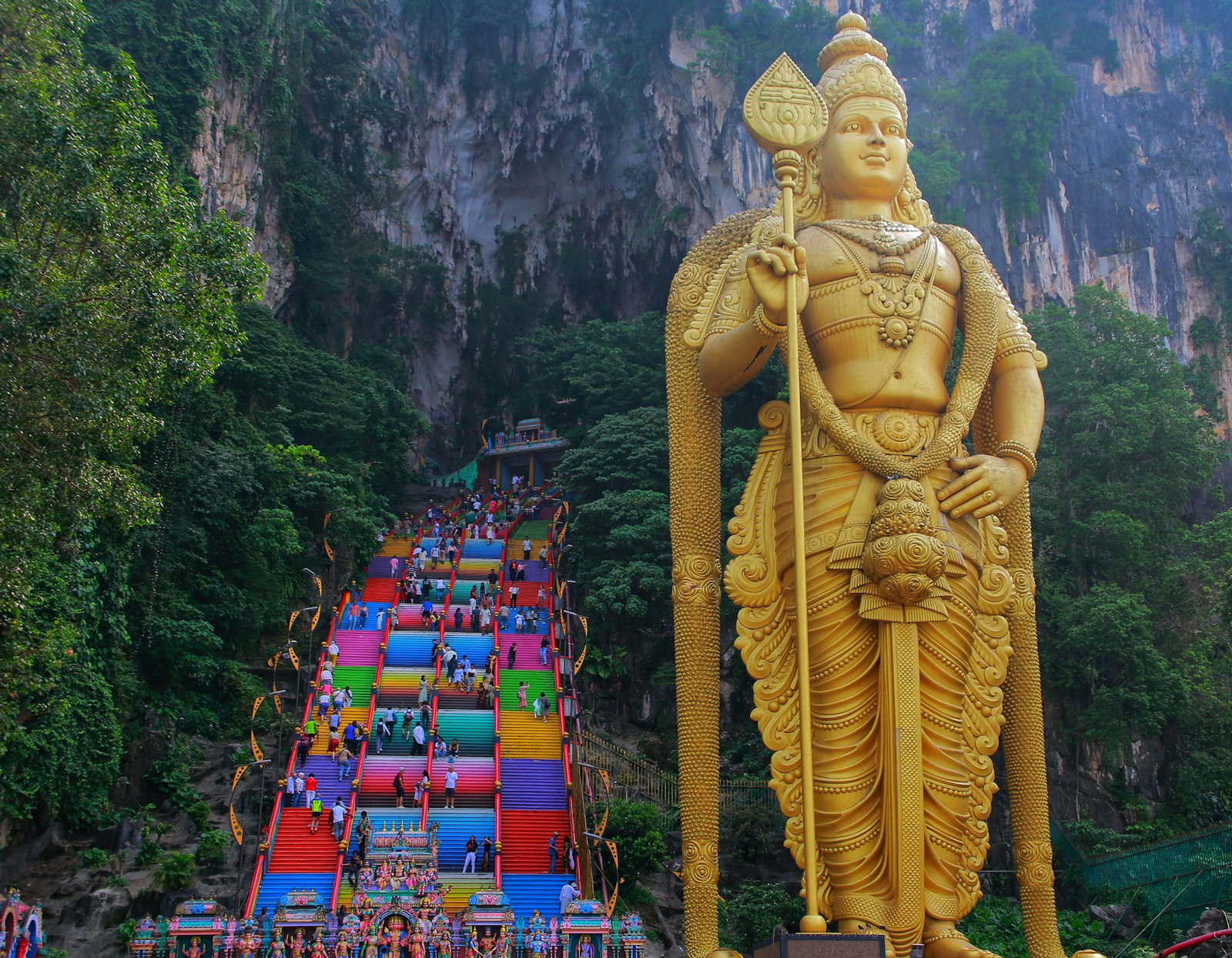 Bright rainbow steps leading up to limestone cave surrounded by jungle, with giant golden statue of Hindu deity standing