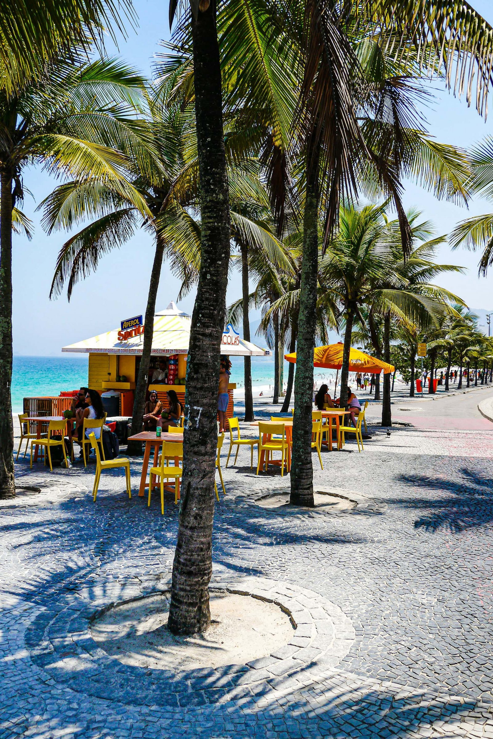 Palm trees by the beach with outdoor vendor and tables with some people sat enjoying the ocean views and chatting