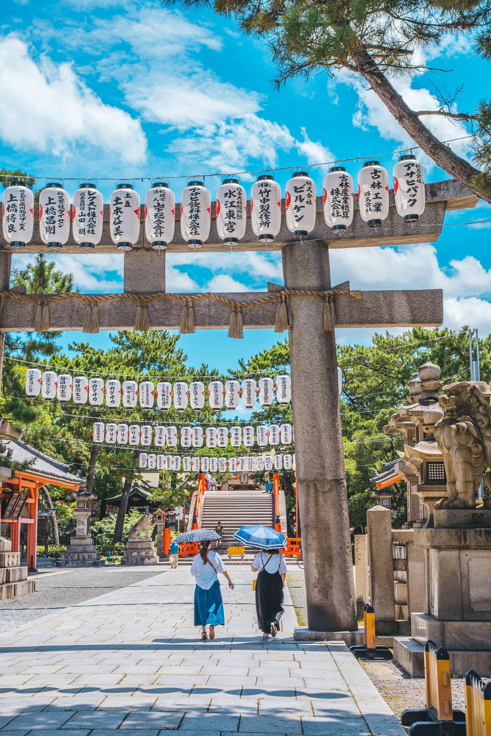 Stone Torii gates with lanterns , stone statuesand bright sky with green trees, two women carrying umbrellas 