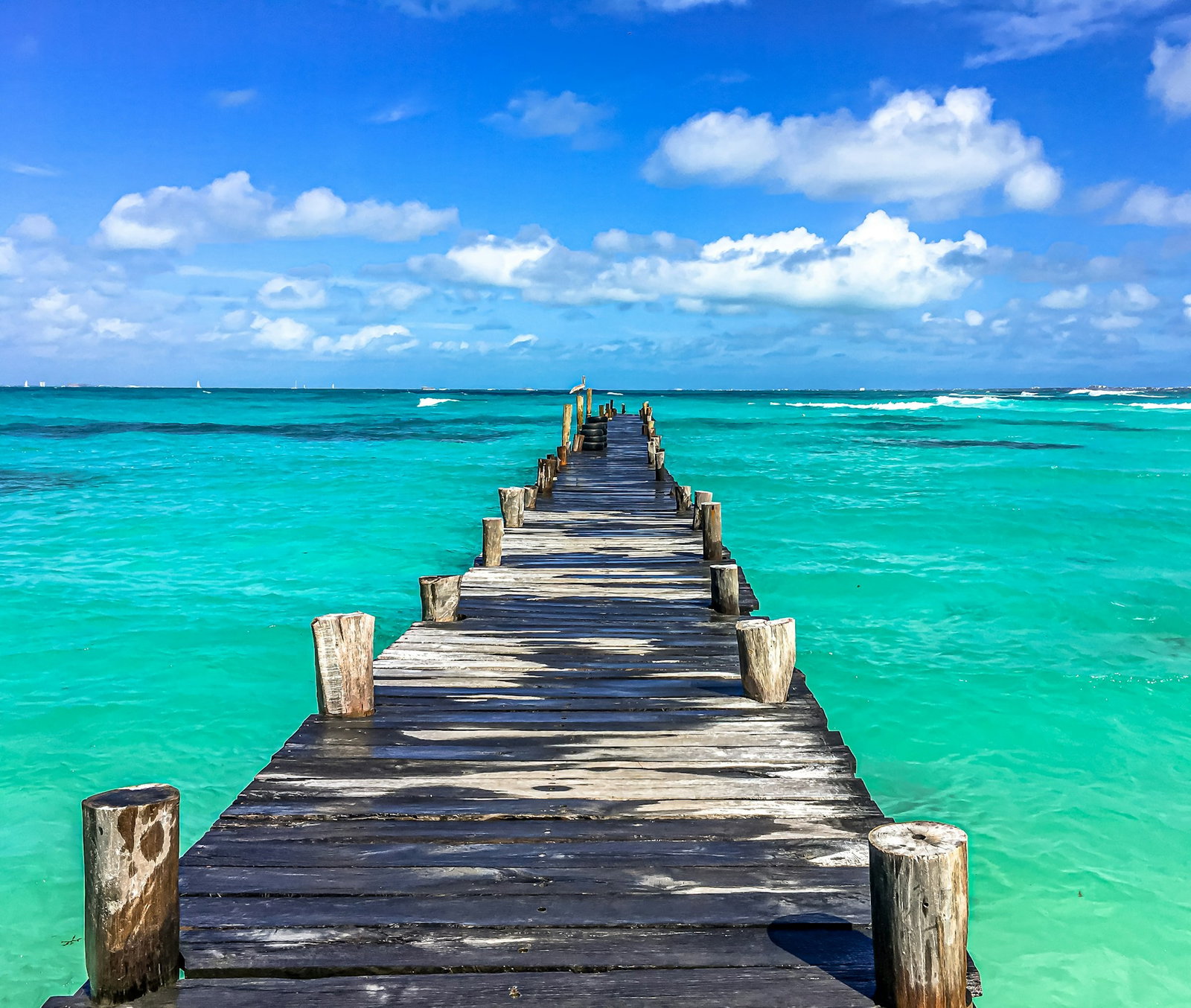 Rustic boardwalk pier leading out to bright turquoise ocean on sunny day with some clouds