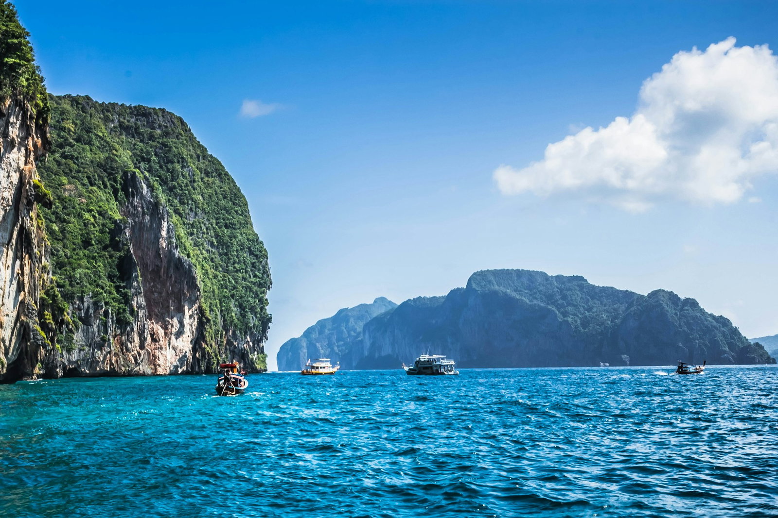 Ocean with limestone boulders in the background covered in lush vegetation, sailboats in the water