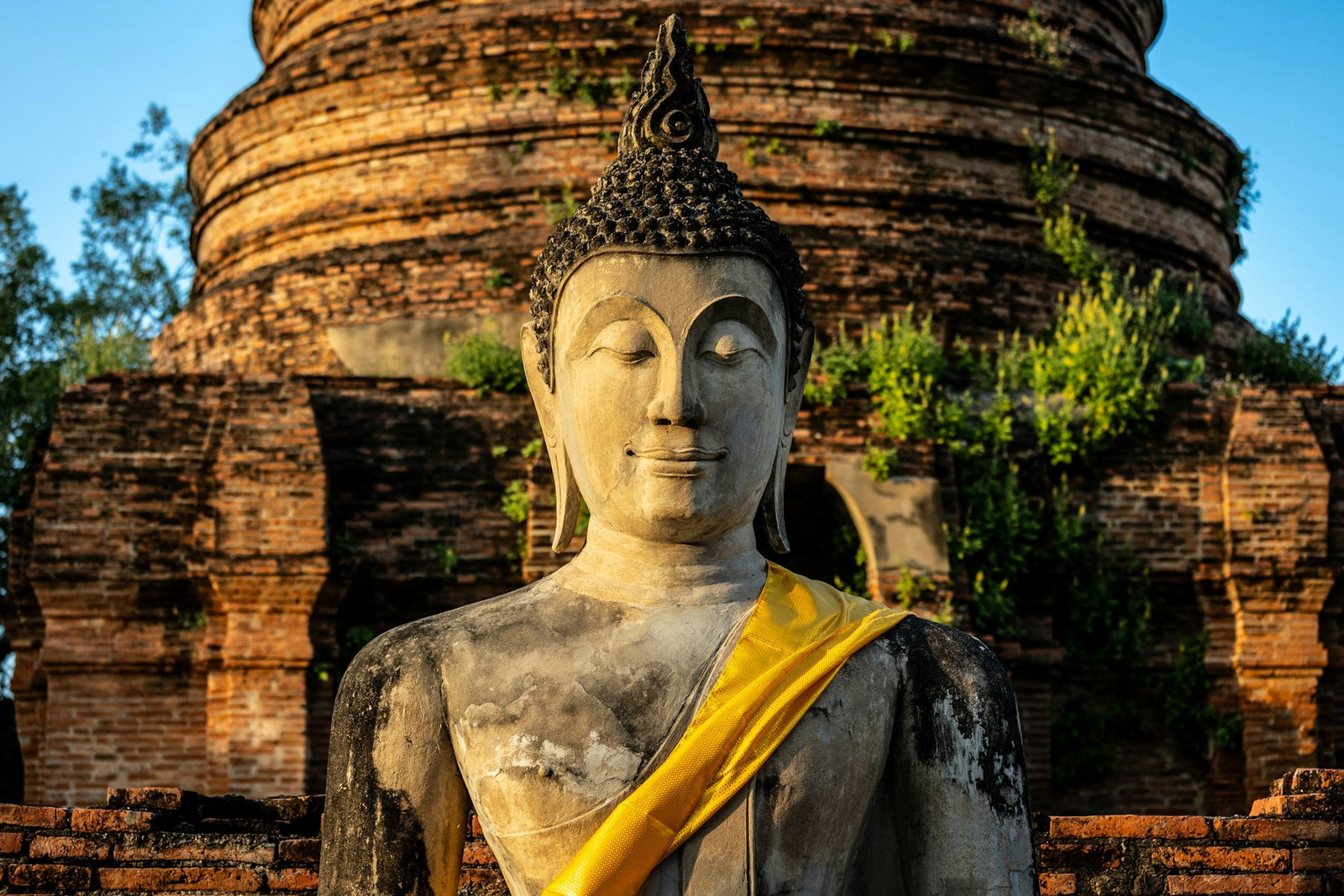 Up-close shot of Buddha statue with yellow silk scarf. Behind statue is a red brick curved temple and a blue sky