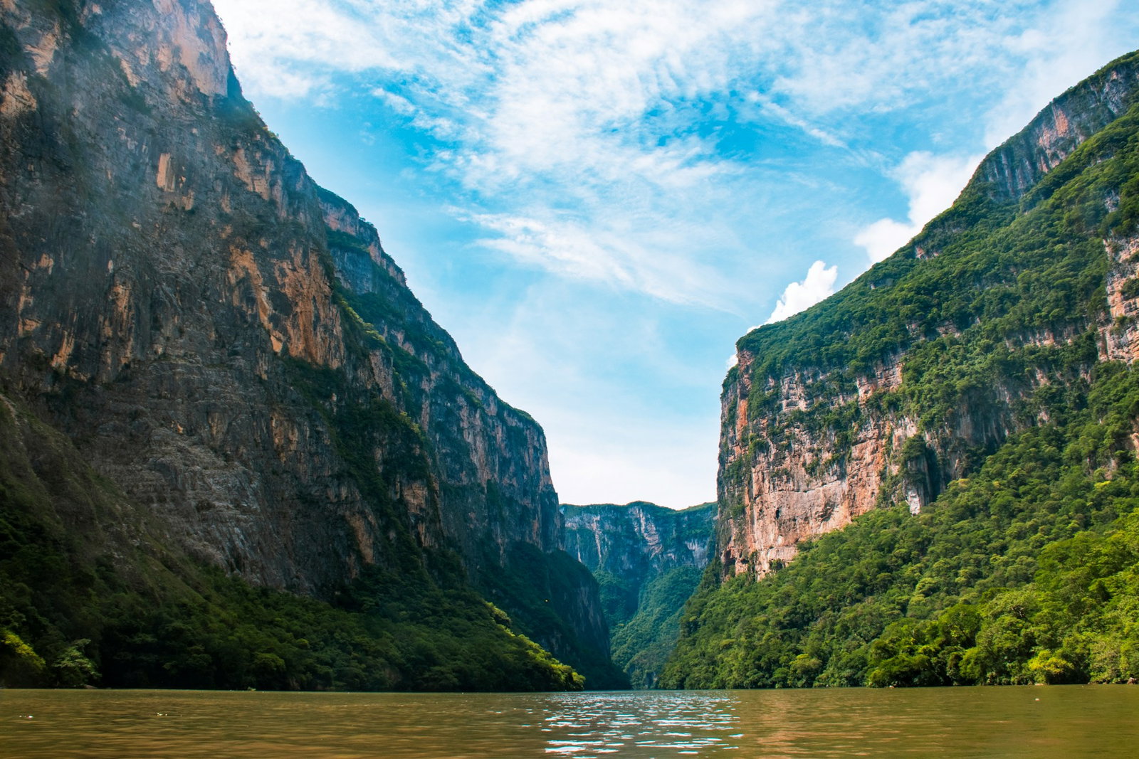 Canyon with giant rocky hills covered in lush green jungle and dark green lake with cloudy blue skies