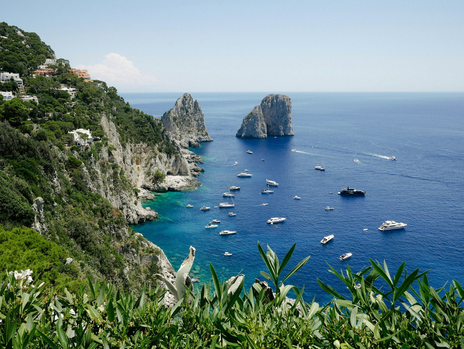 Green shubbery and grey cliffs with villas built in to the side overlooking sapphire ocean with small cruise ships