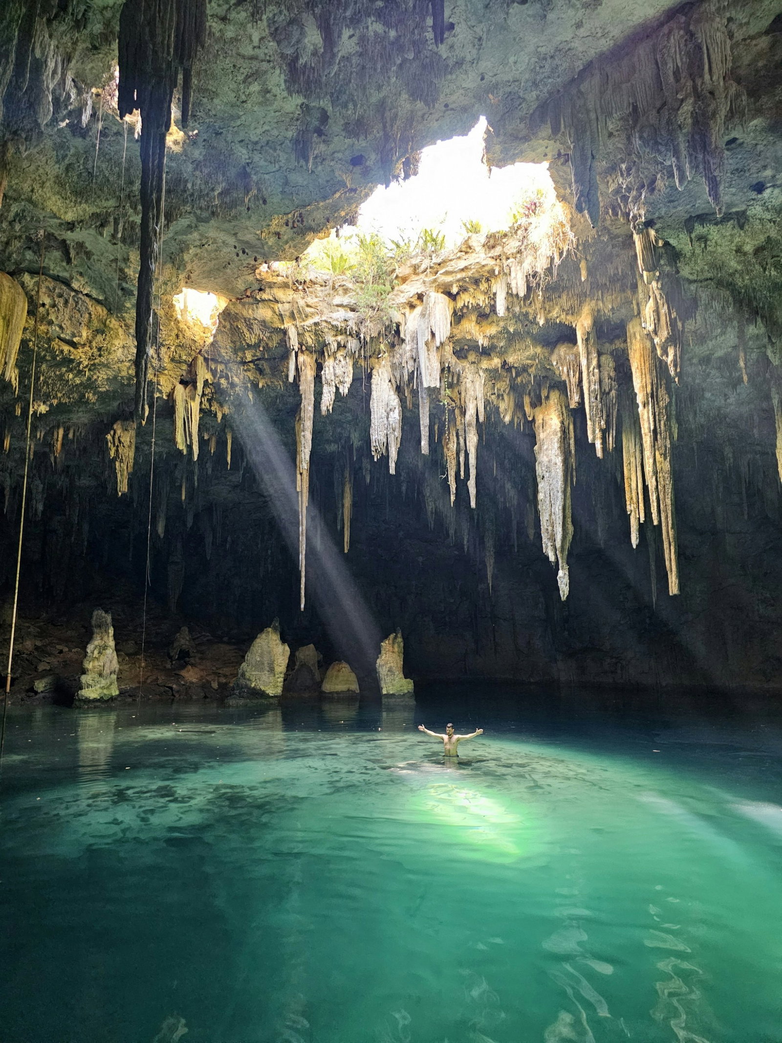 Secret cave pool with sunlight pouring from the centre over a person with their arms out in bright turquoise waters