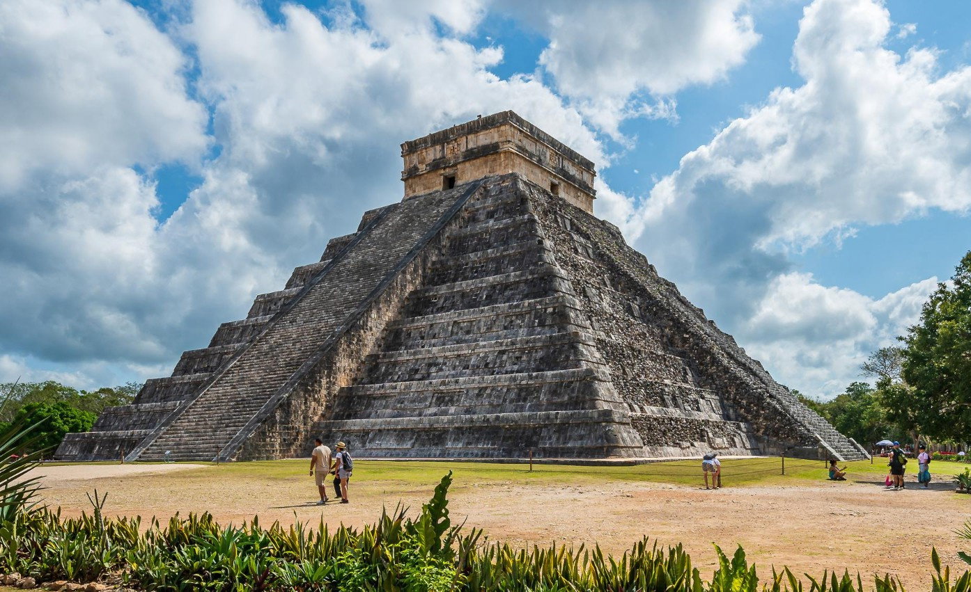 Ancient pyramid with stairs facing out onto emerald field with tourists exploring on a cloud day