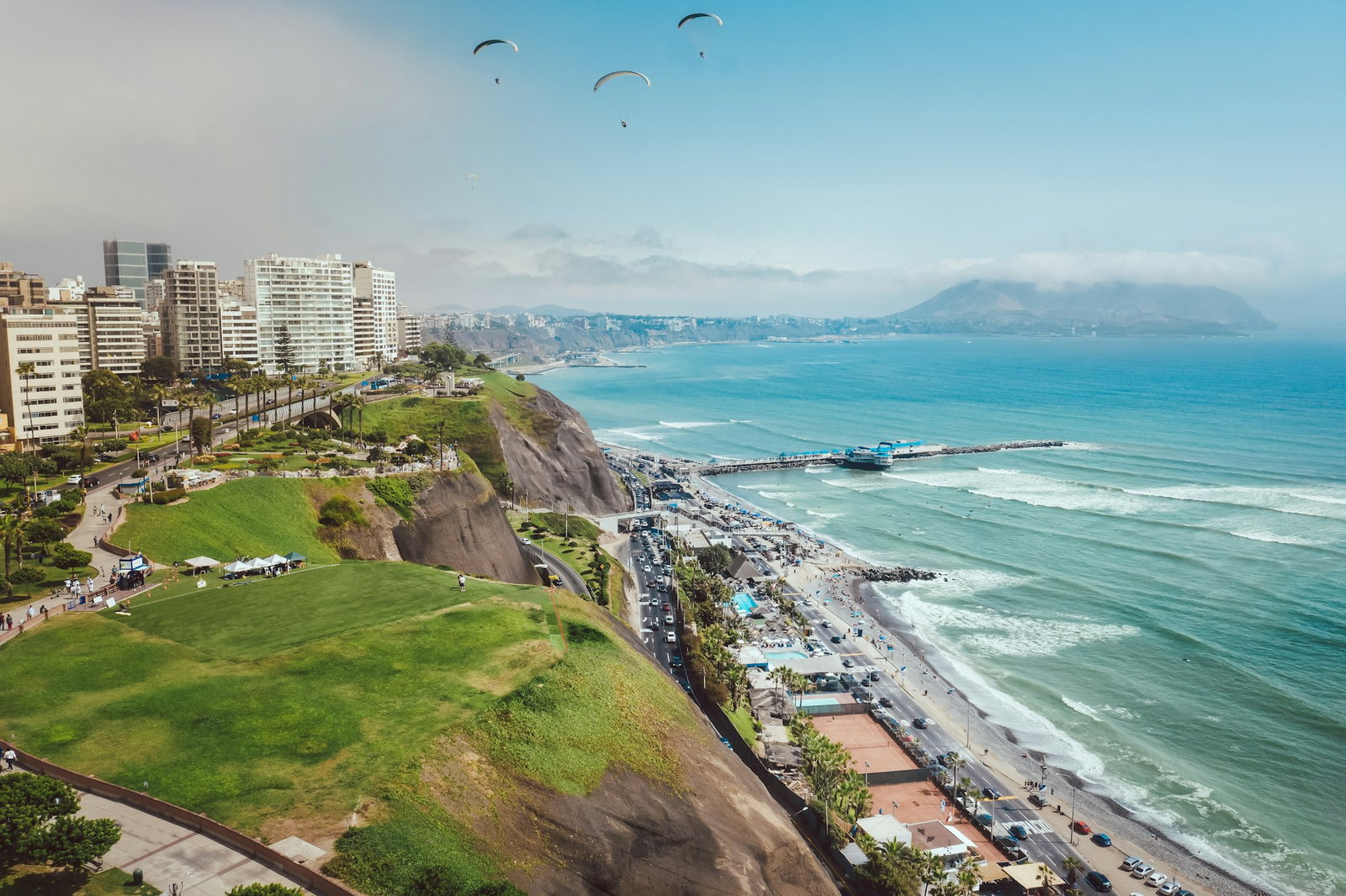 Modern city with green hills and road right by the coast, with paragliders flying through the blue sky