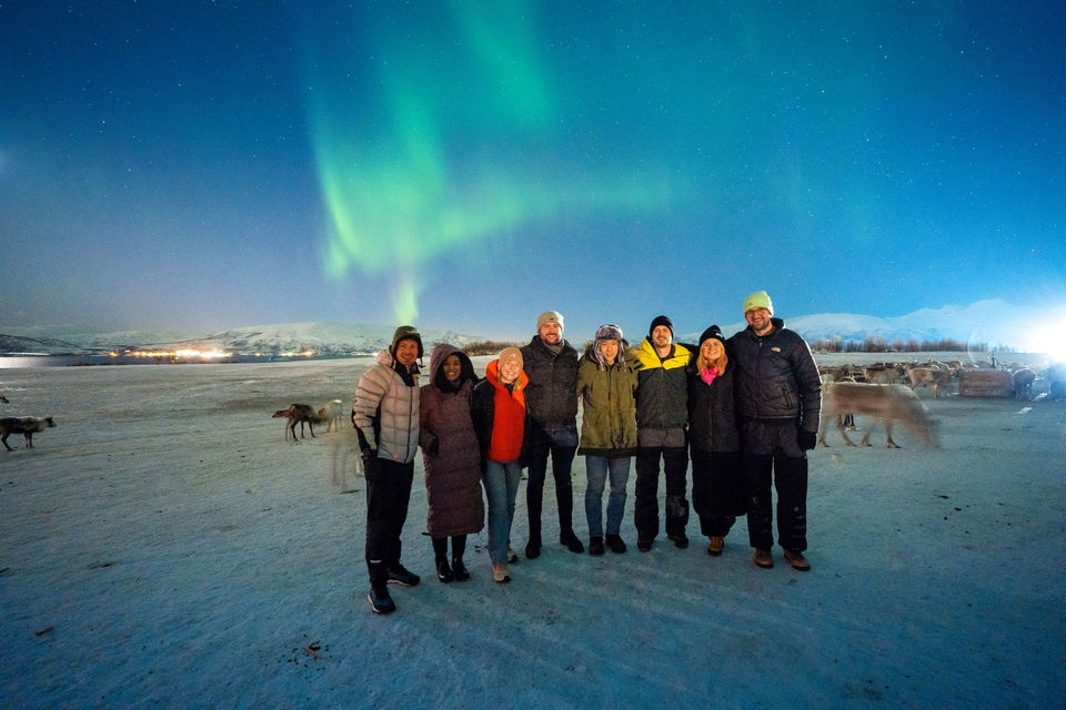 Group of travellers in Norway, standing in the snow next to reindeer and Northern lights