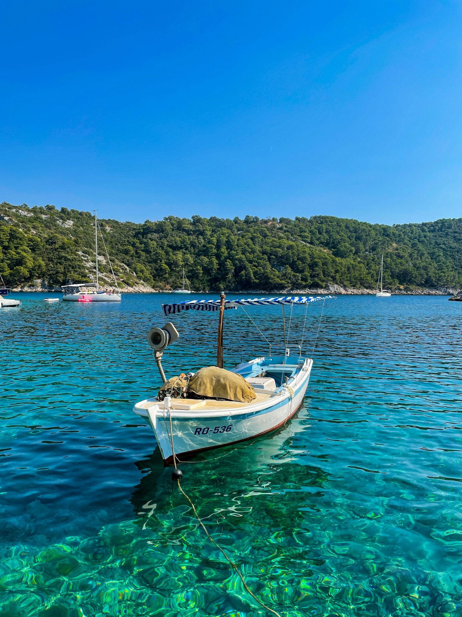 Small canoe boat docked in bright sapphire water with sailboat in distance and lush hills