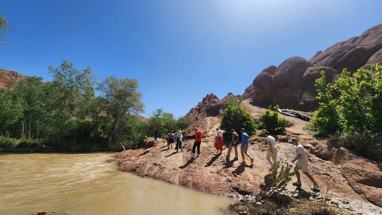People walking along the river inside rugged orange canyon under bright blue sky