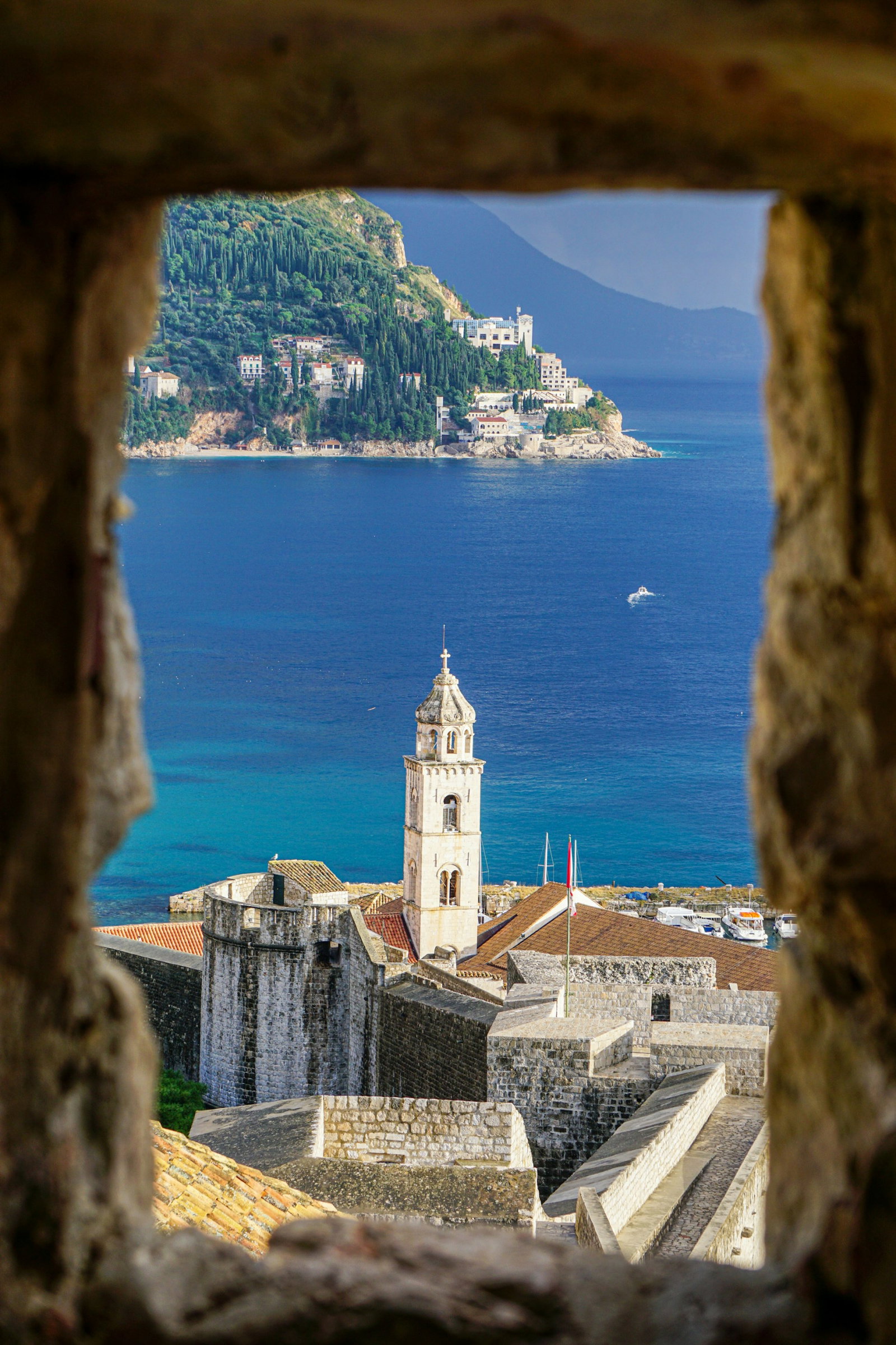 City wall window peering out onto Dubrovnik's Medieval architecture and blue ocean with hilly island in near distance