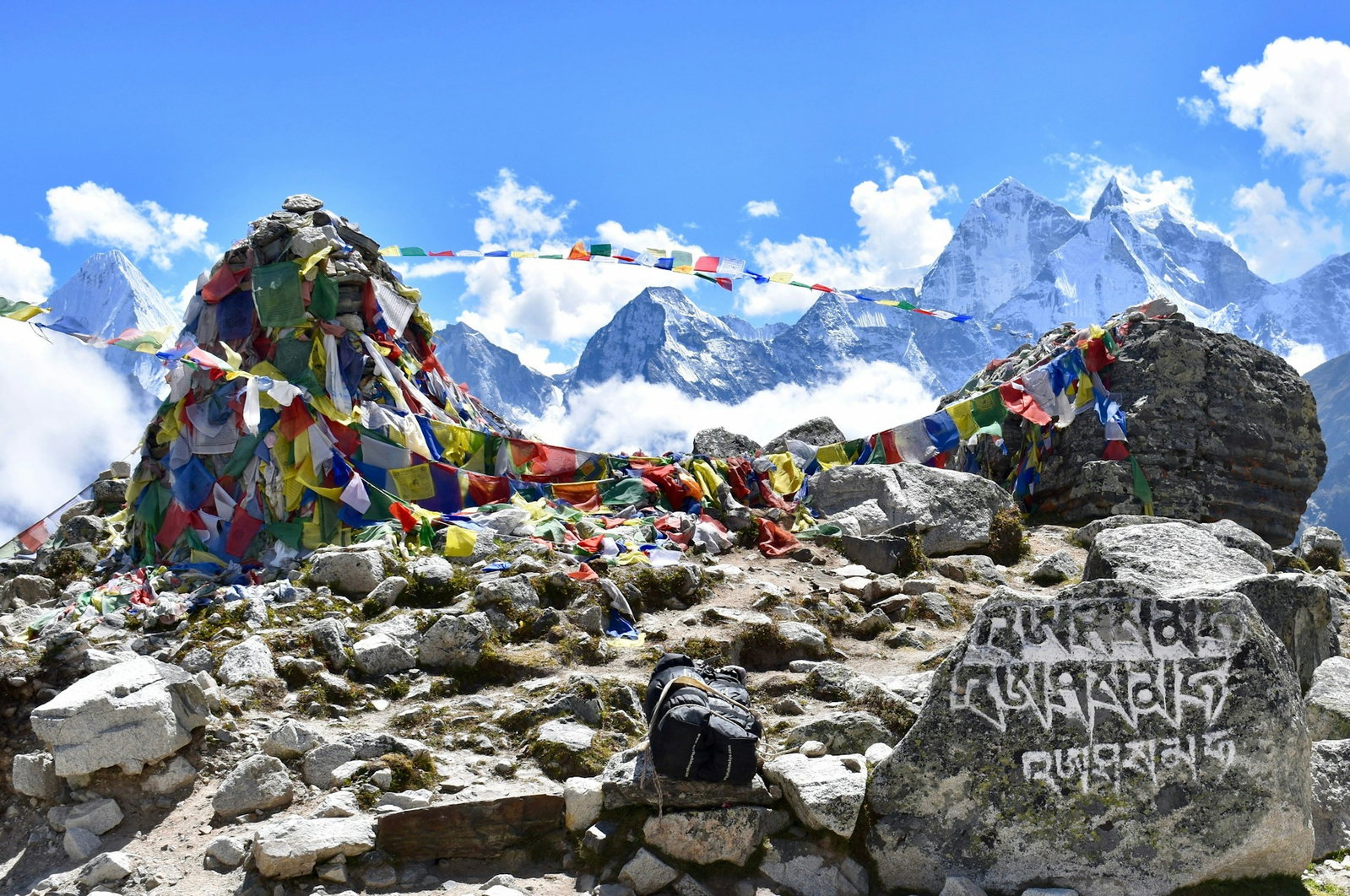 Grey rocks with rainbow coloured flags tied around the base and overlooking snowy mountains with white clouds and blue sky