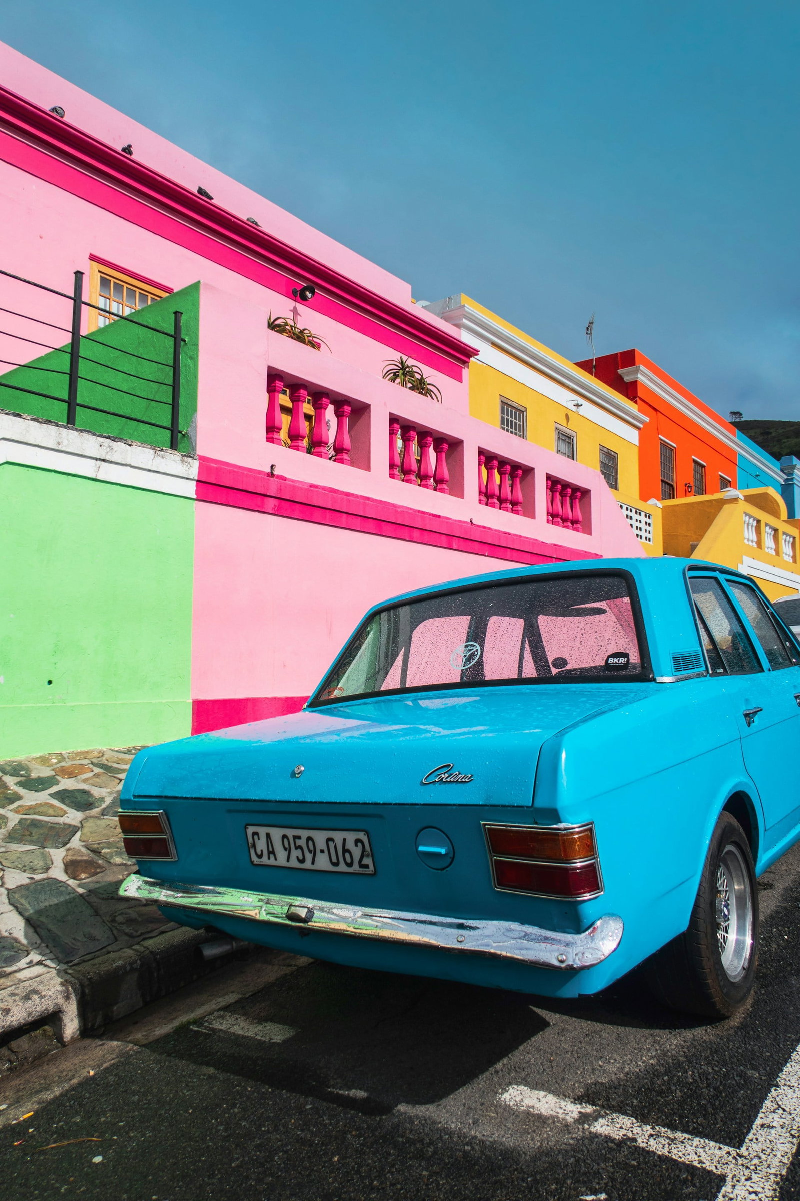 Vintage blue car parked in front of row of brightly painted houses, green, pink, yellow, orange and blue on a sunny day