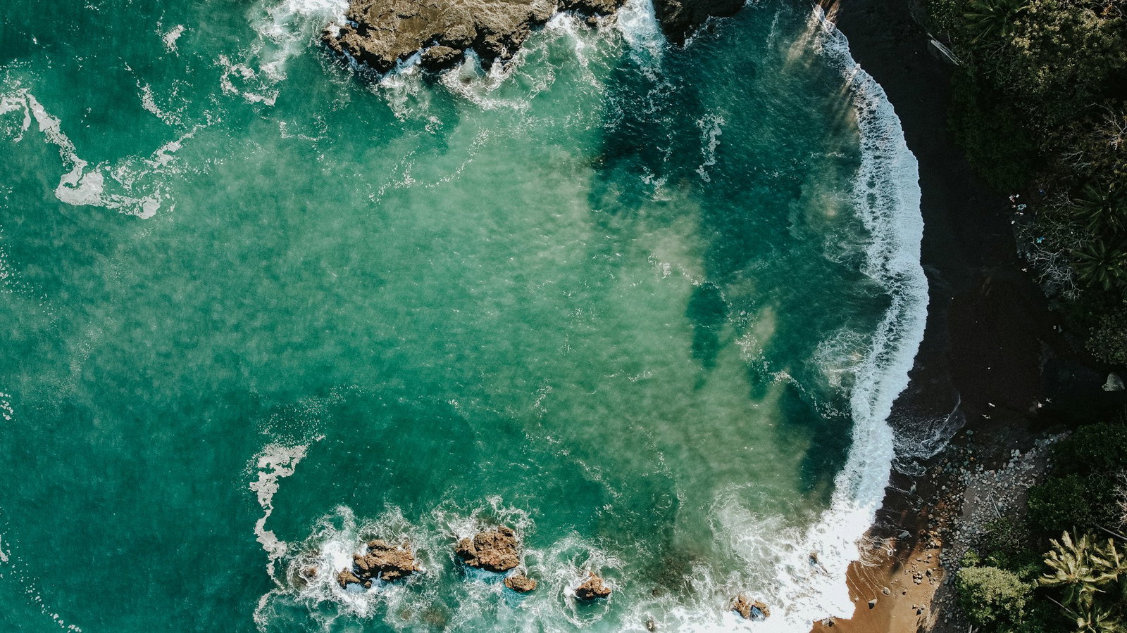 Aerial shot of teal ocean with crashing waves against black sand and charcoal-coloured rocks, glimpse of jungle to the right