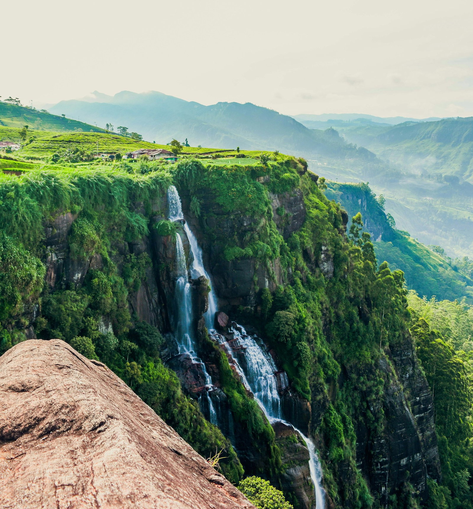 Cascading waterfall falling from cliff covered in lush vegetation with more valleys in the distance, taken from a red rock