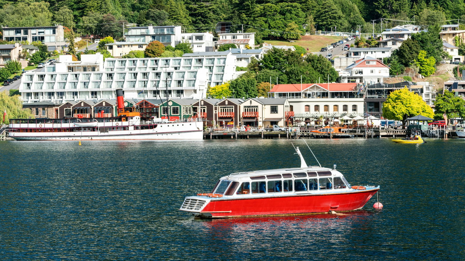 Red canal cruise on water with buildings in distance inside harbour city of Queenstown