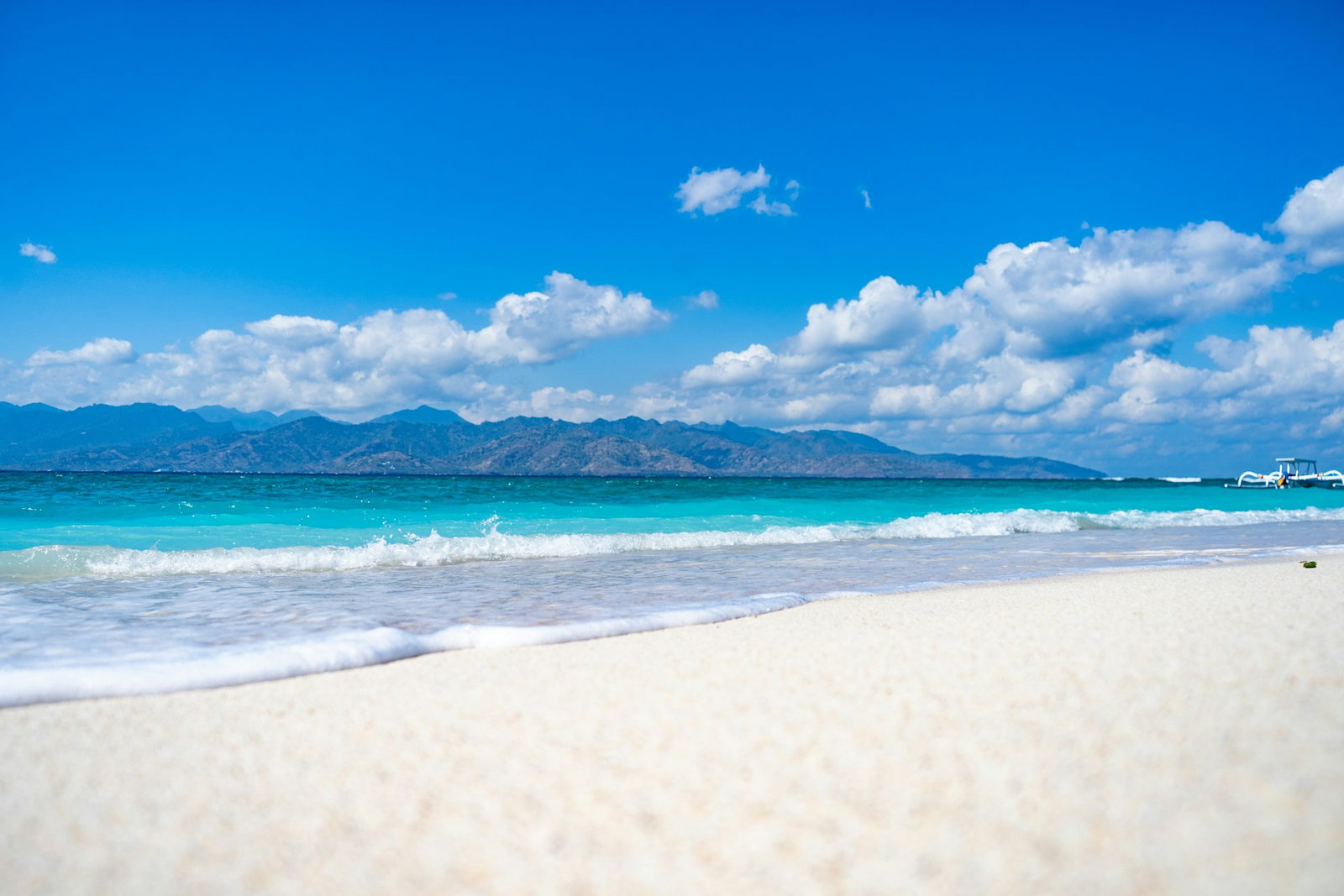 White sandy beach with bright turquoise ocean and a boat to the right, and an island in the distance, Gili Trawangan