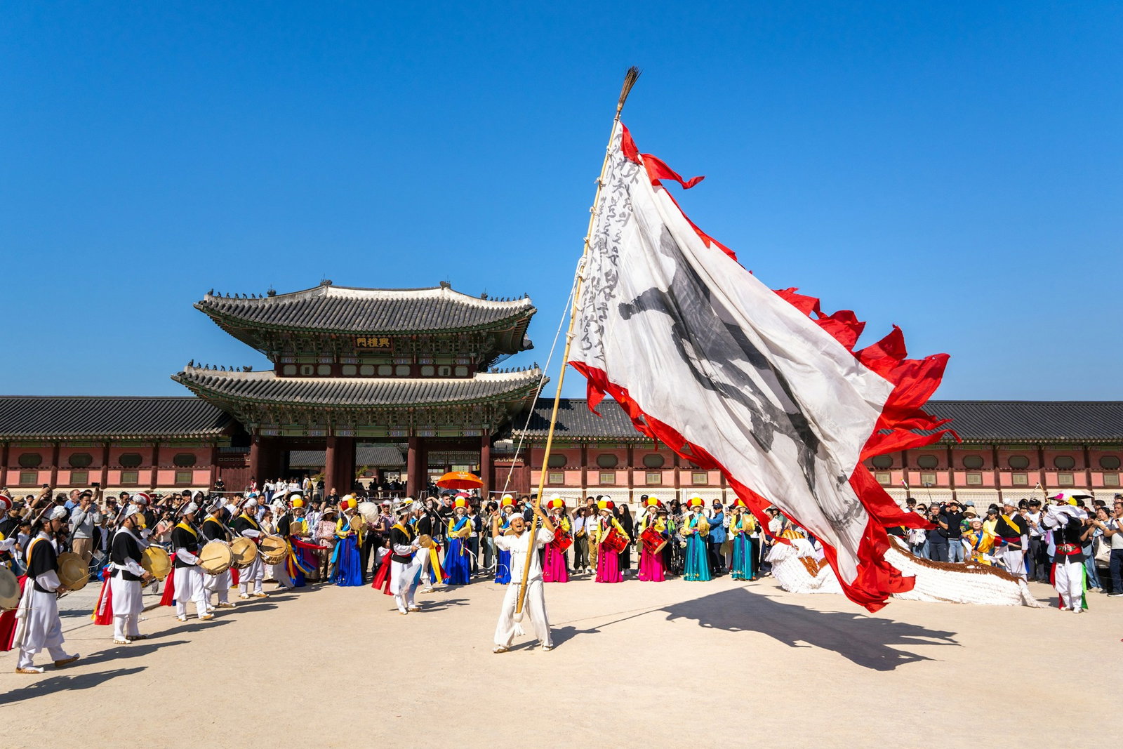 Person raising a giant flag surrounded by band playign traditional instruments and crowd of tourists outside palace