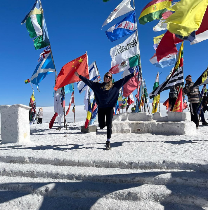 Traveller at the Uyuni Salt Flats with their hands in the air next to the different world flags.