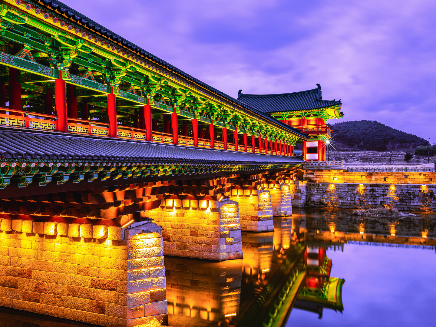 Illuminated traditional bridge with bright red columns and emerald details with pagoda rooftops under violet sky and waters