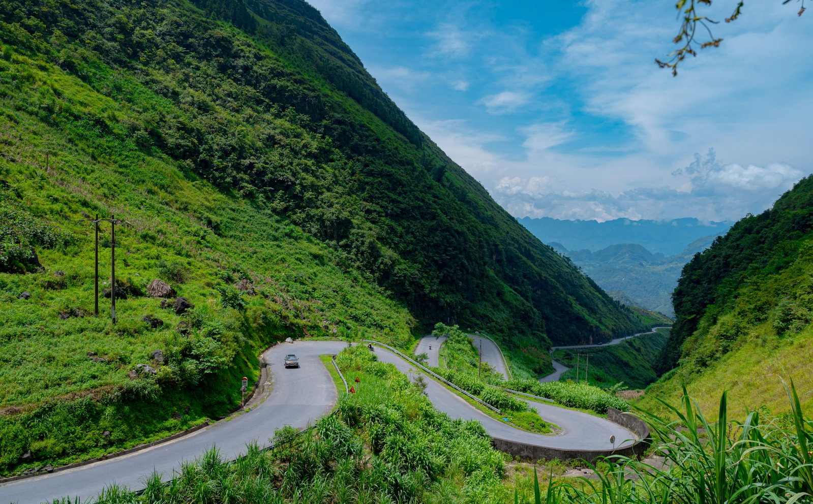 Winding grey road in lush valley filled with emerald shrubbery and valley hills with cloudy blue sky in the distance