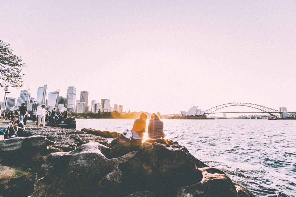 Two travellers sitting on rocks by the water with Sydney Harbour Bridge in the background