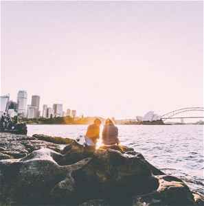Two travellers sitting on rocks by the water with Sydney Harbour Bridge in the background