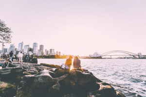 Two travellers sitting on rocks by the water with Sydney Harbour Bridge in the background