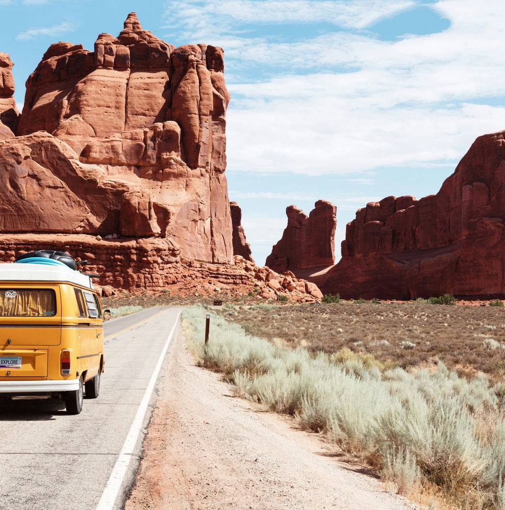 A lone yellow campervan drives in a road between canyons in the USA