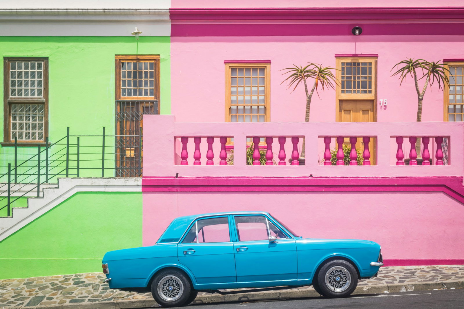Old blue car parked in front of bright pink and lime green houses with bannisters and wooden doors, Cape Town