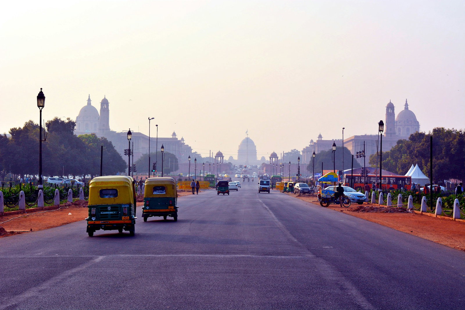 Tuk-tuk cars driving through wide street with orange sand and buildings, New Delhi