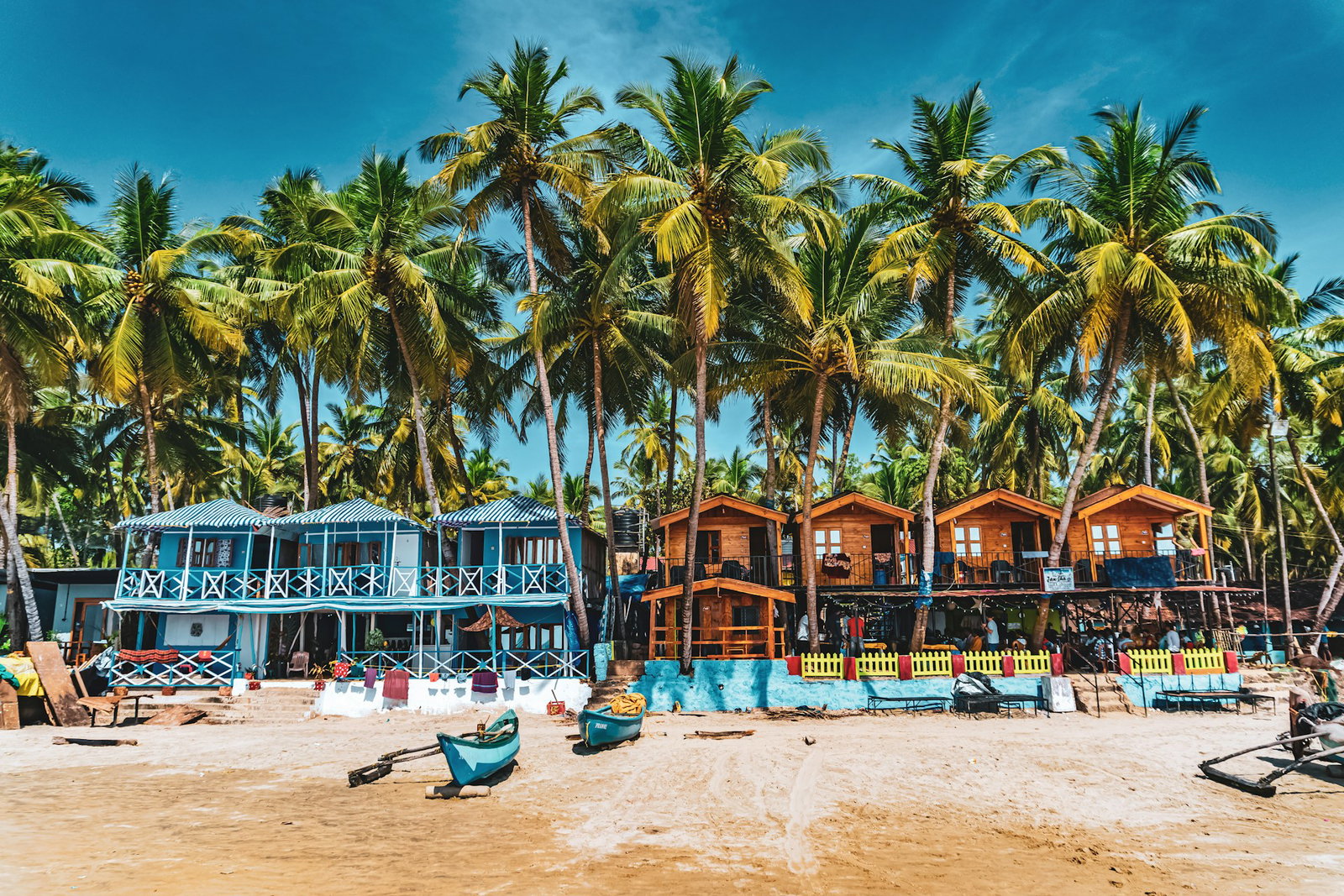 Rows of blue and brown beach houses with palm trees in between on a golden beach with boats, Goa