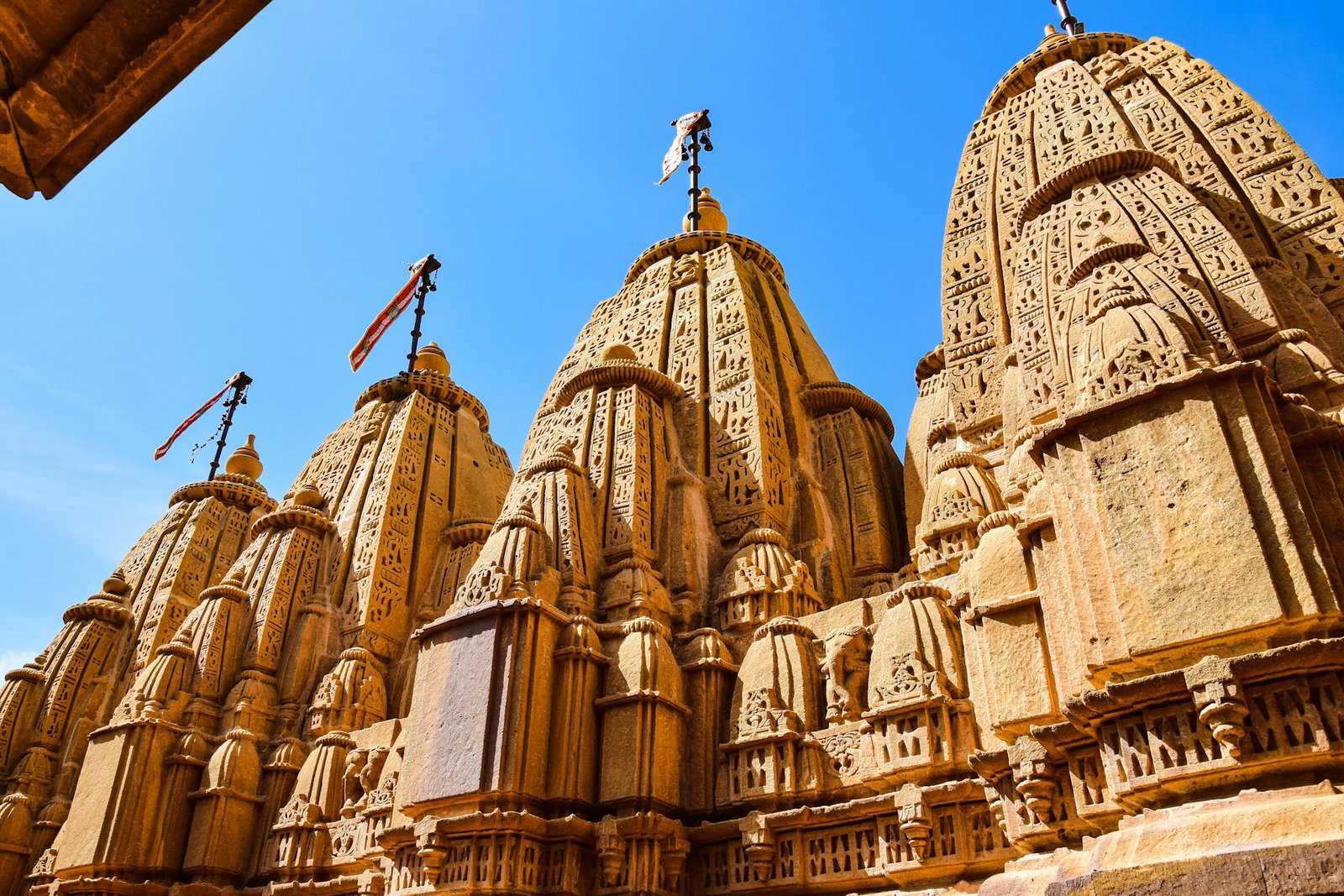 Golden, sandstone temples carved with intricate mosaics and curved roofs with a small pole and flag on top, Jaisalmer