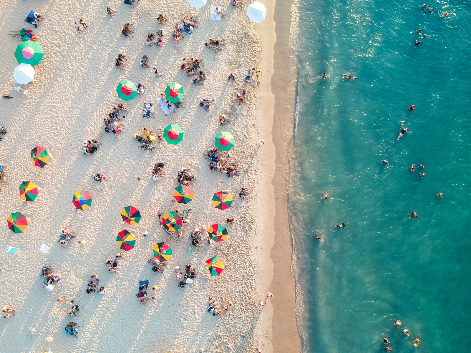 Birdseye shot of a white sand beach with colourful umbrellas and sunbathers with bright water that has swimmers in it
