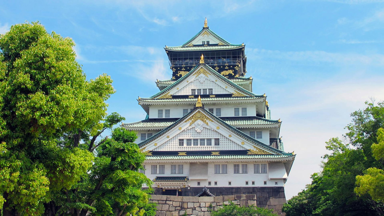 Blue sky over a white traditional Japanese castle with green roofs and gold details, next to two bright green trees.