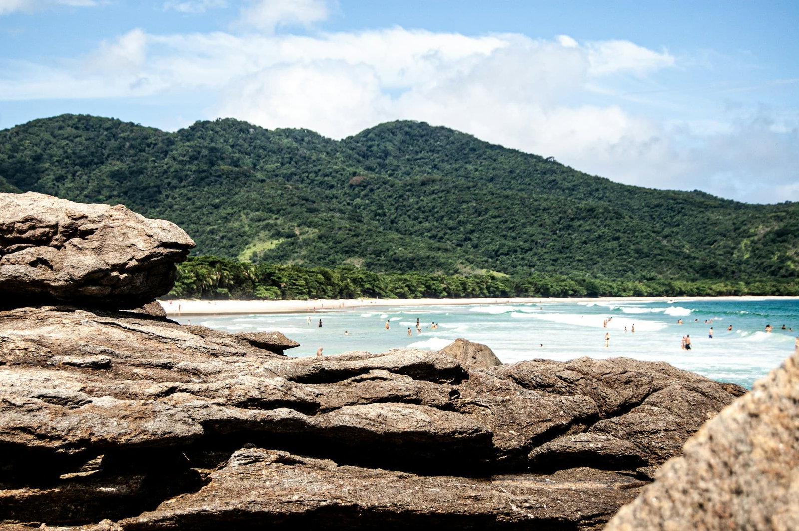 Rock with beach hidden behind it and people swimming with jungle hills in the background