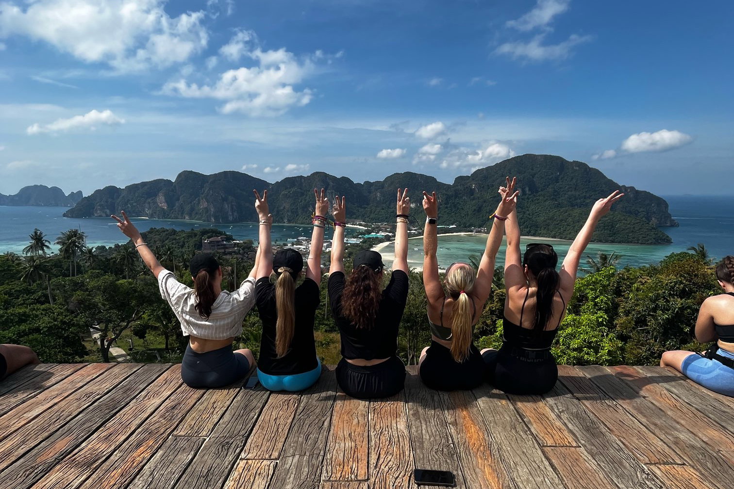 A group of travellers sit on a platform overlooking the ocean and islands while doing peace hand signs 