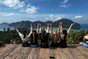 A group of travellers sit on a platform overlooking the ocean and islands while doing peace hand signs 