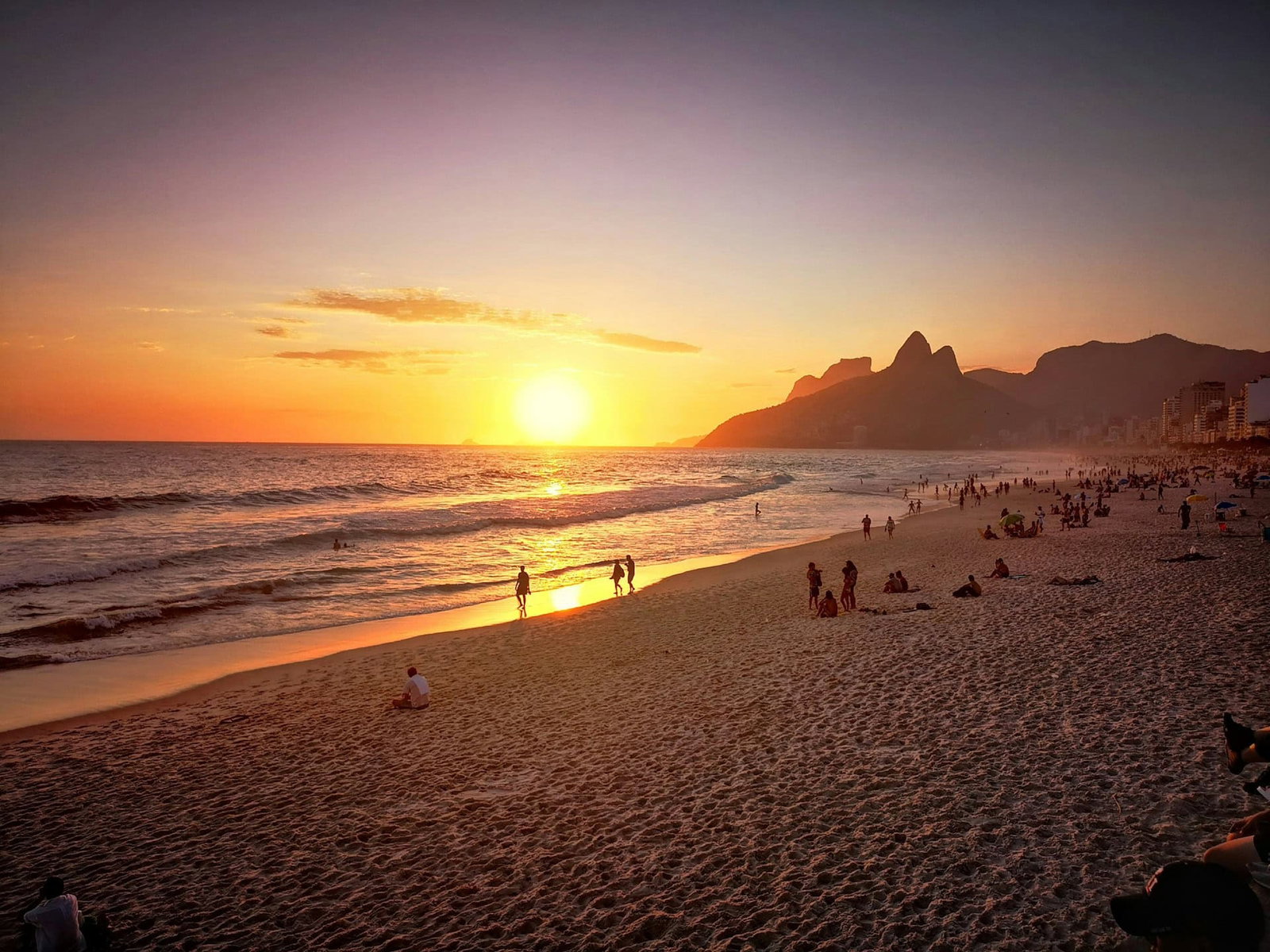 Sunset by golden beach with people hanging out and walking along the ocean with mountains and skyscrapers in background
