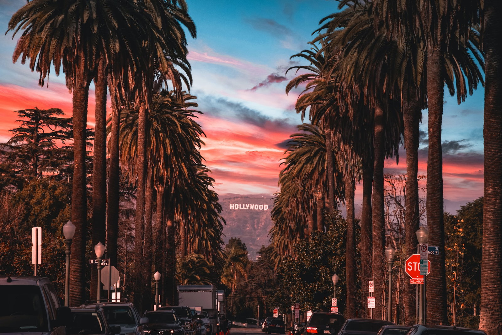 A palm tree-lined street at sunset with hills and the Hollywood sign in the background