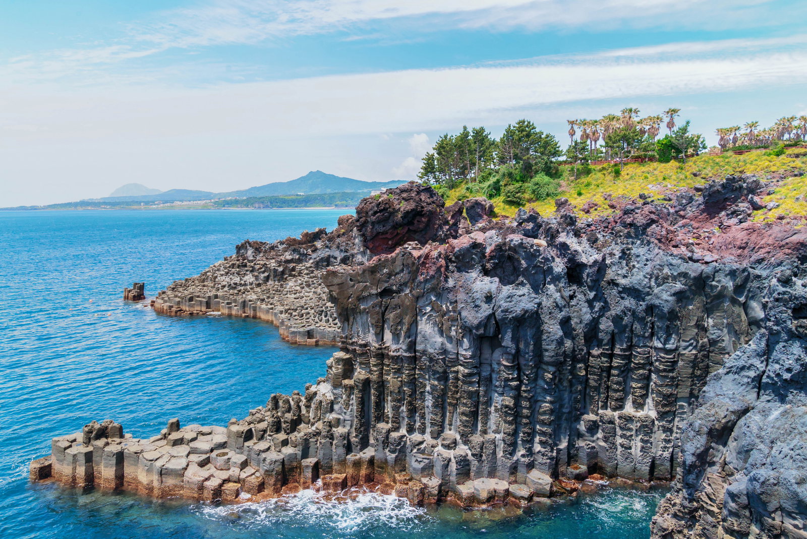 Unique grey rock formations by cliff edge with emerald flora and sapphire sea with volcano backdrop and blue skies