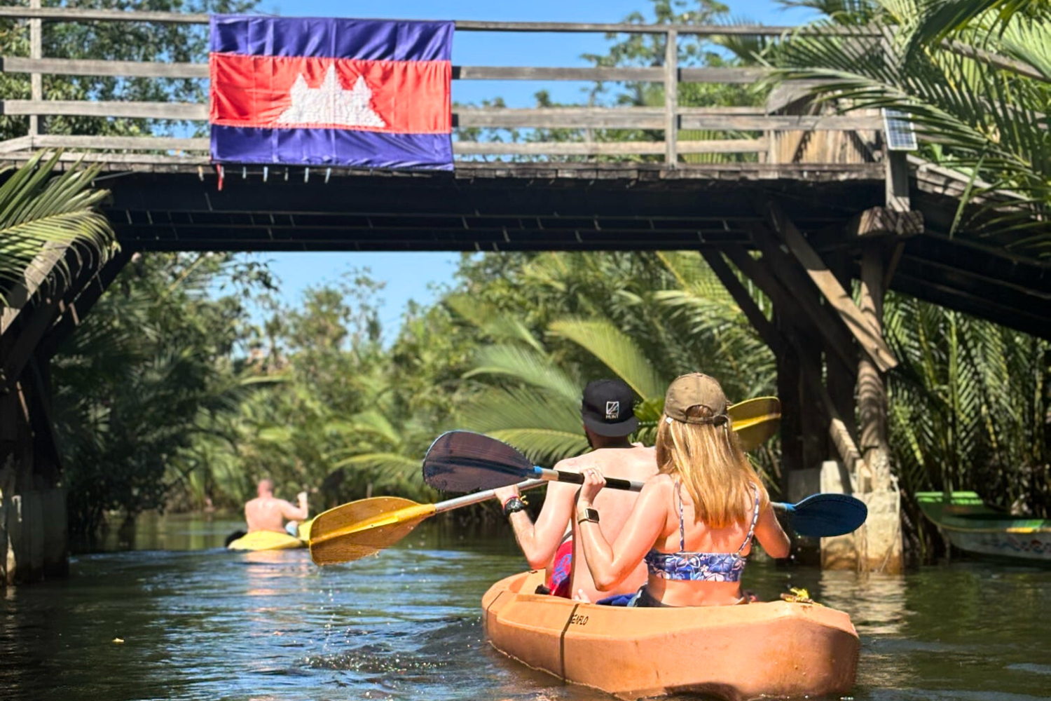 Travellers kayaking through river in Kampot, under a bridge with Cambodia flag